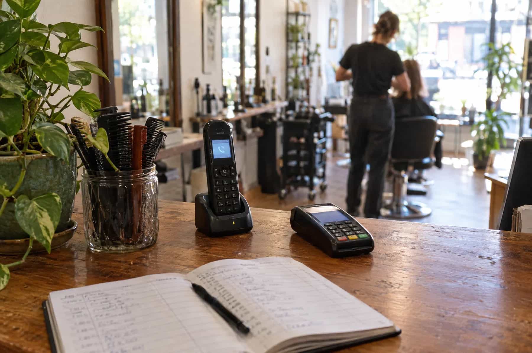 A cordless phone ringing in its cradle on an empty hair salon counter, with a stylist at work in the background