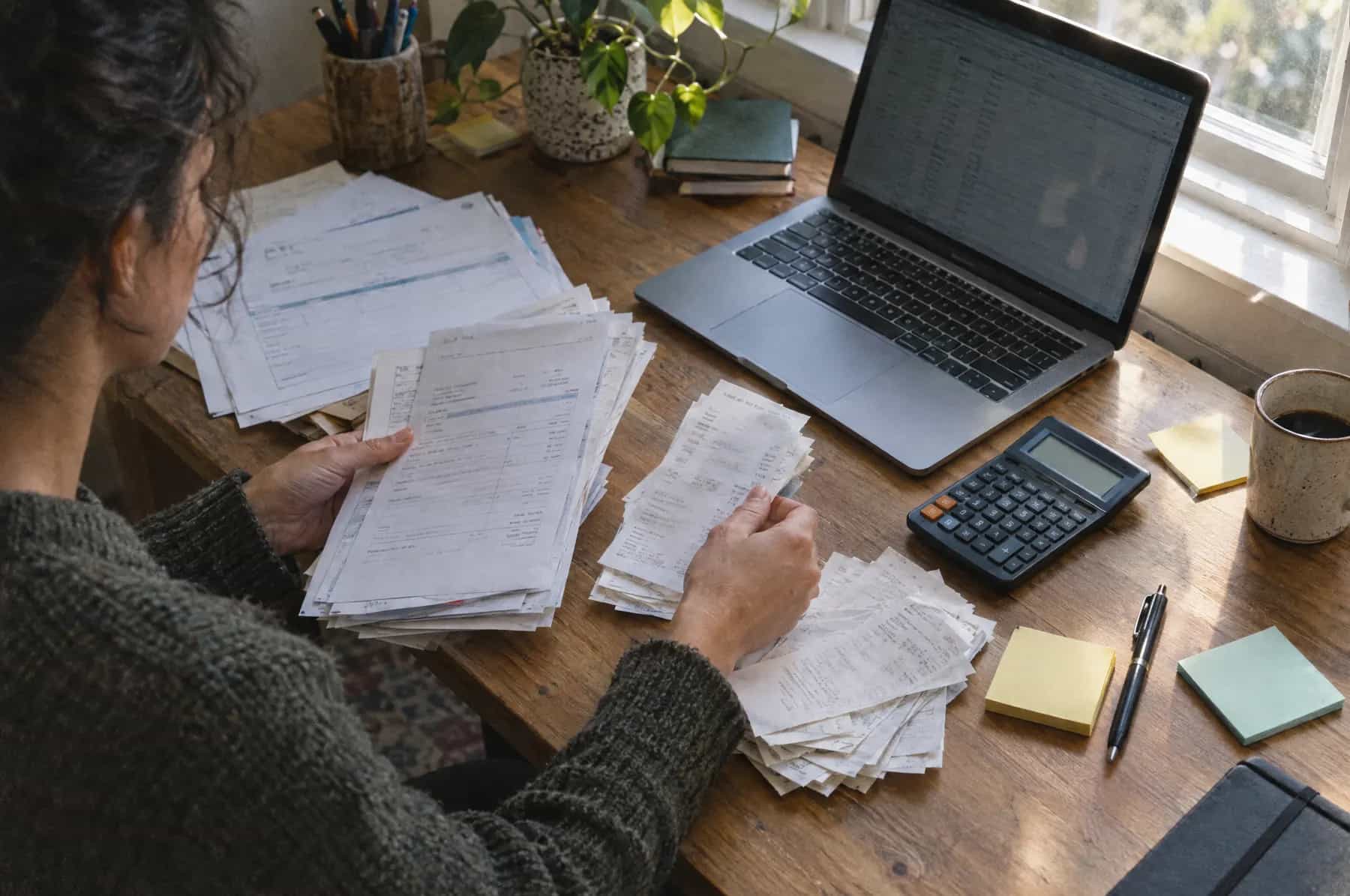 A business owner sorting paper invoices and receipts beside a laptop and calculator